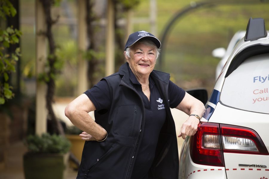 A woman with white hair in an RFDS hat and vest leans against an RFDS car, smiling into the camera.